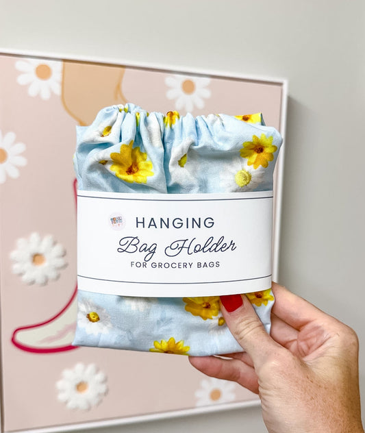 A hand holds a light blue fabric hanging bag holder with a yellow sunflower print and a white paper label that reads 'Hanging Bag Holder for Grocery Bags.' The background shows a beige wall with framed floral artwork.