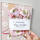 A hand holds a pink floral hanging bag holder with a white paper label that reads 'HANGING Bag Holder FOR GROCERY BAGS.' The fabric has a strawberry and leaf print.