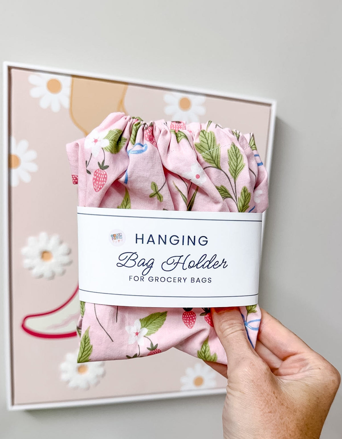 A hand holds a pink floral hanging bag holder with a white paper label that reads 'HANGING Bag Holder FOR GROCERY BAGS.' The fabric has a strawberry and leaf print.