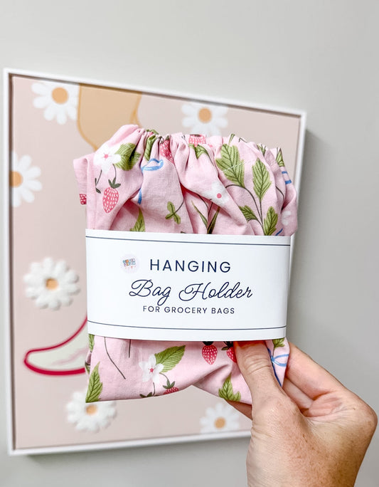 A hand holds a pink floral hanging bag holder with a white paper label that reads 'HANGING Bag Holder FOR GROCERY BAGS.' The fabric has a strawberry and leaf print.