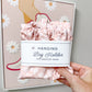 A hand holds a pink floral hanging bag holder wrapped in a white paper band labeled 'Hanging Bag Holder FOR GROCERY BAGS'. The background shows a framed art print against a neutral wall.