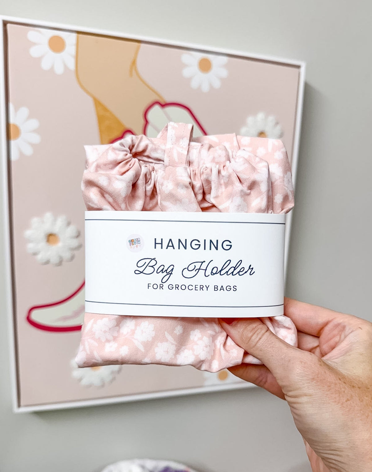 A hand holds a pink floral hanging bag holder wrapped in a white paper band labeled 'Hanging Bag Holder FOR GROCERY BAGS'. The background shows a framed art print against a neutral wall.