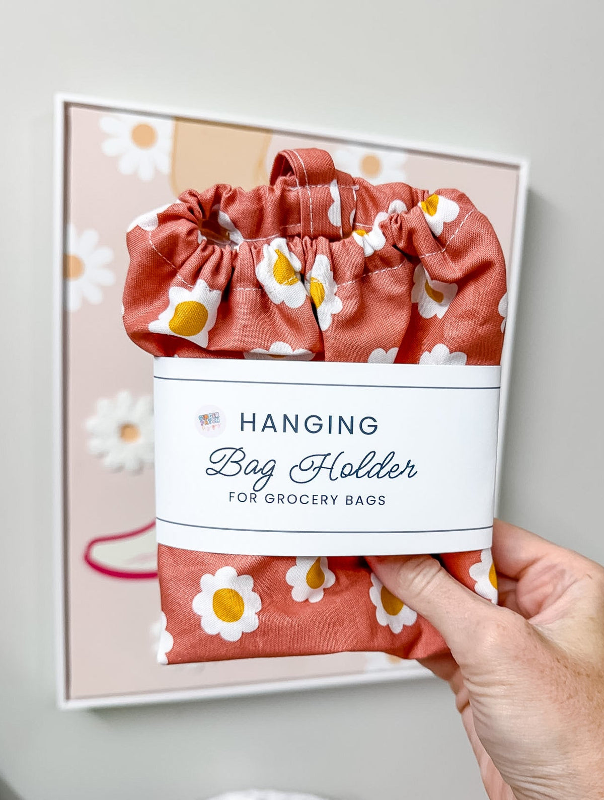 A hand holds a terracotta-colored fabric hanging bag holder with a white daisy print and a white paper label that reads 'Hanging Bag Holder for Grocery Bags.' The background shows a framed matching bag holder on a light gray wall.