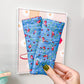 A hand with olive green nail polish holds up a pair of blue quilted wrist warmers featuring a red and green floral pattern, set against a home office background with a framed print on the wall.