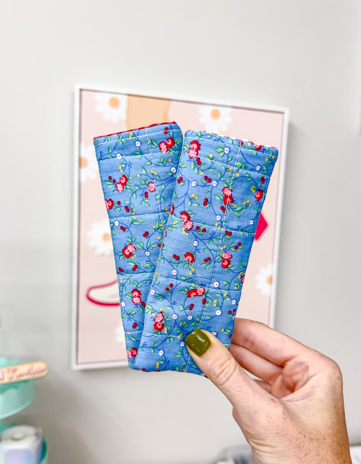 A hand with olive green nail polish holds up a pair of blue quilted wrist warmers featuring a red and green floral pattern, set against a home office background with a framed print on the wall.