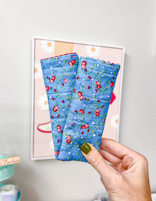 A hand with olive green nail polish holds up a pair of blue quilted wrist warmers featuring a red and green floral pattern, set against a home office background with a framed print on the wall.