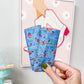 A hand with olive green nail polish holds up a small, quilted fabric cup in a blue floral print. The background shows a light gray wall with a framed art print and a mint green decorative stand on a wooden table.