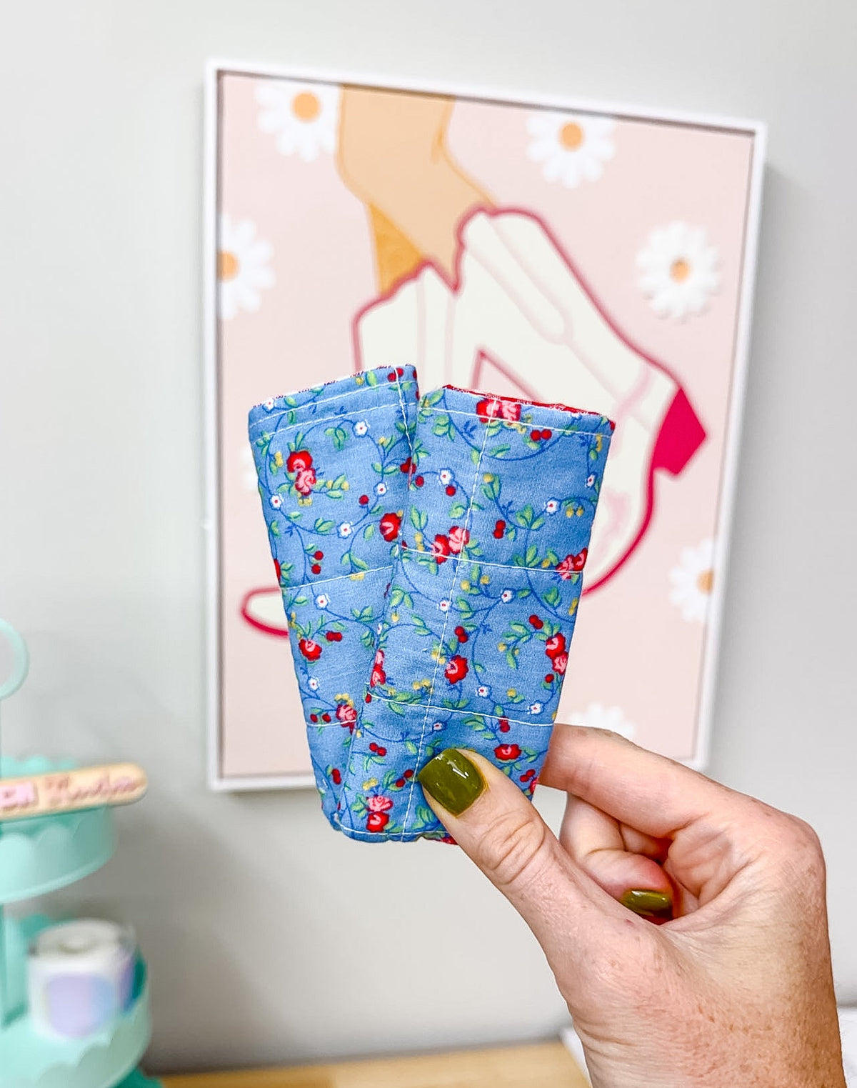 A hand with olive green nail polish holds up a small, quilted fabric cup in a blue floral print. The background shows a light gray wall with a framed art print and a mint green decorative stand on a wooden table.