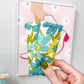 A hand with olive green nail polish holds a small fabric pouch decorated with a teal, yellow, and green floral pattern on a white background. The hand is positioned in front of a light gray wall with a framed floral print.
