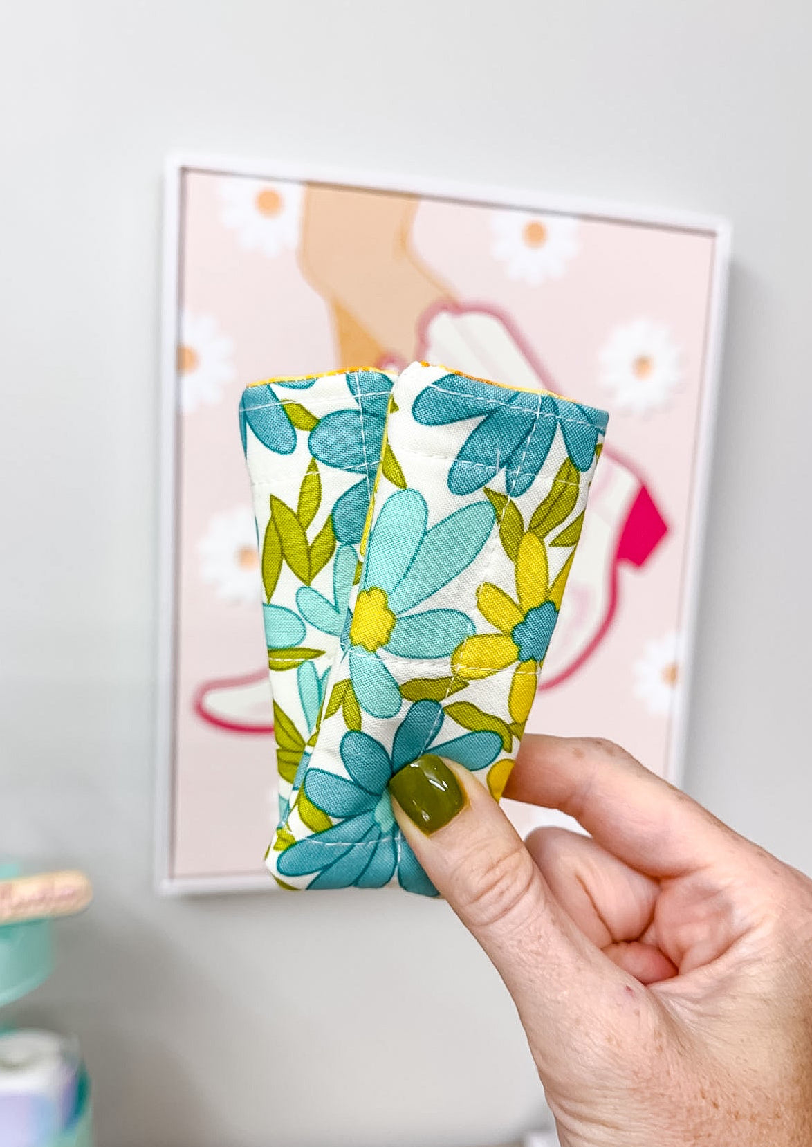 A hand with olive green nail polish holds a small fabric pouch decorated with a teal, yellow, and green floral pattern on a white background. The hand is positioned in front of a light gray wall with a framed floral print.