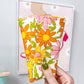 A hand with olive green nail polish holds a small fabric pouch or case featuring a retro floral pattern of orange, pink, and yellow flowers on a white background. The background shows a light gray wall with a framed picture and a wooden desk.