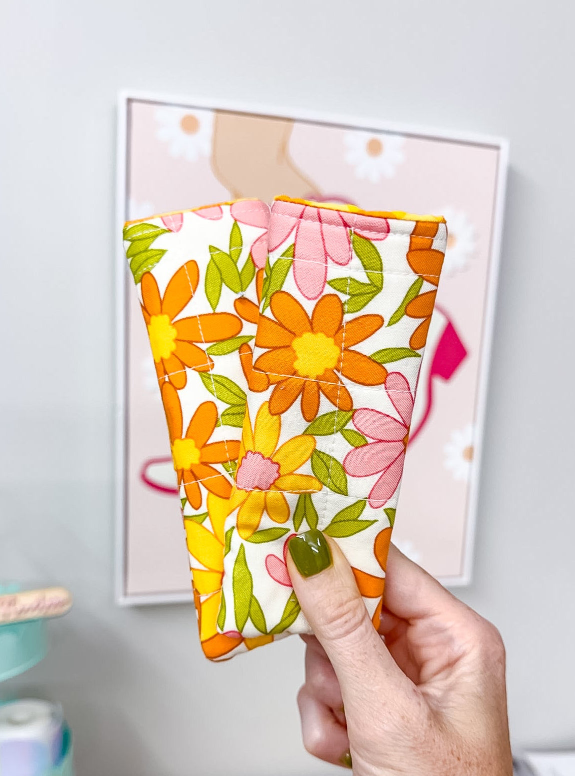 A hand with olive green nail polish holds a small fabric pouch or case featuring a retro floral pattern of orange, pink, and yellow flowers on a white background. The background shows a light gray wall with a framed picture and a wooden desk.