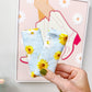 A hand holds two light blue fabric items with a yellow and white daisy floral print in front of a framed art print on a light gray wall.