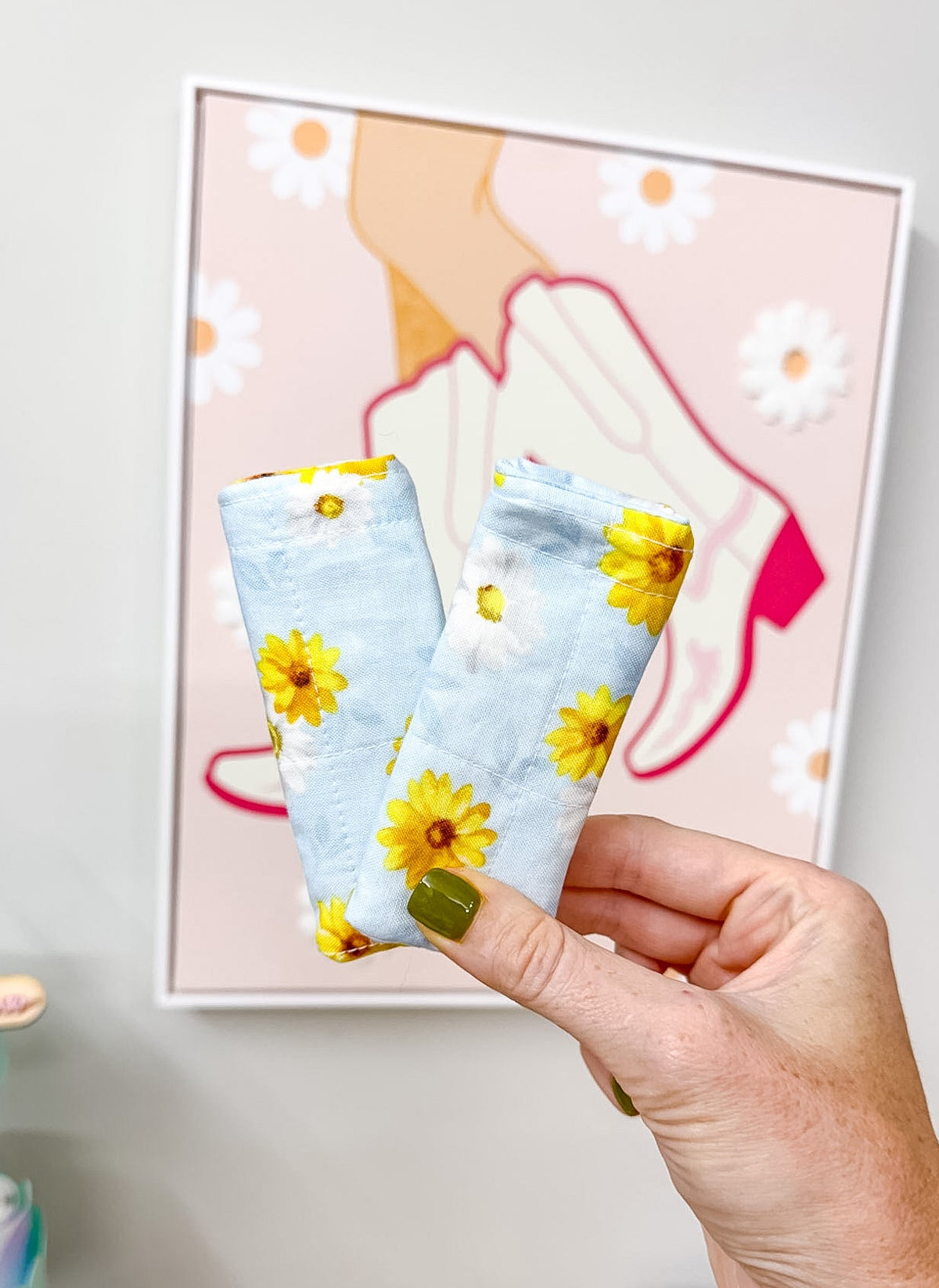 A hand holds two light blue fabric items with a yellow and white daisy floral print in front of a framed art print on a light gray wall.