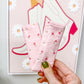 A hand with olive green nail polish holds up a pair of light pink ankle socks with a small red and white floral pattern, set against a background of a framed art print and a wooden table.