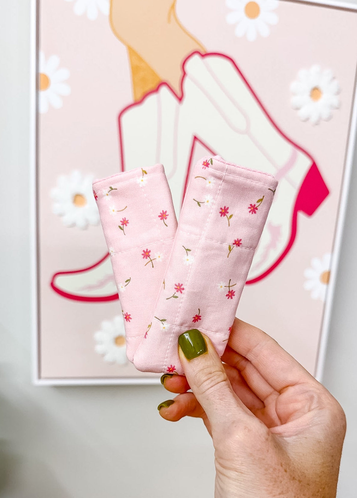 A hand with olive green nail polish holds up a pair of light pink ankle socks with a small red and white floral pattern, set against a background of a framed art print and a wooden table.