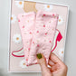 A hand with green nail polish holds a pair of light pink fabric leg warmers with a small floral print, set against a gray wall with a framed print.