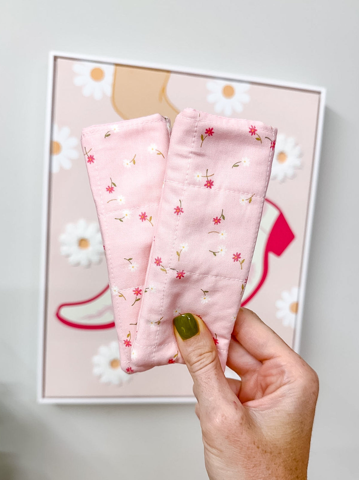 A hand with green nail polish holds a pair of light pink fabric leg warmers with a small floral print, set against a gray wall with a framed print.