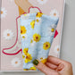 A hand with green nail polish holds a light blue fabric pouch decorated with a yellow and white daisy floral pattern, positioned in front of a framed art print on a gray wall.