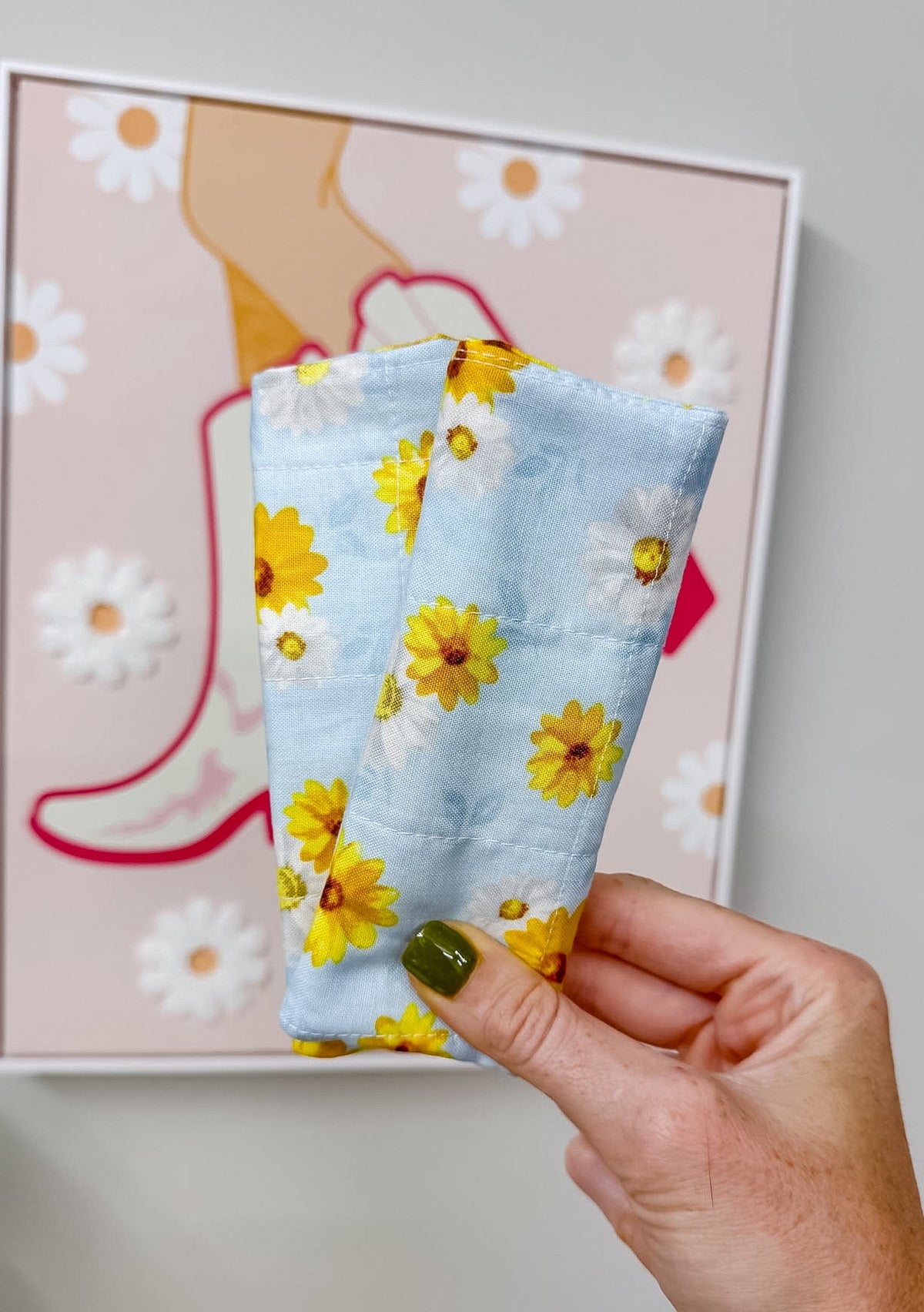 A hand with green nail polish holds a light blue fabric pouch decorated with a yellow and white daisy floral pattern, positioned in front of a framed art print on a gray wall.