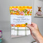 A hand holds a packaged hanging towel set against a gray wall. The set includes a folded floral towel with pink, orange, and yellow flowers, a folded orange-and-white floral towel, and a folded white towel with a sage green plaid pattern.
