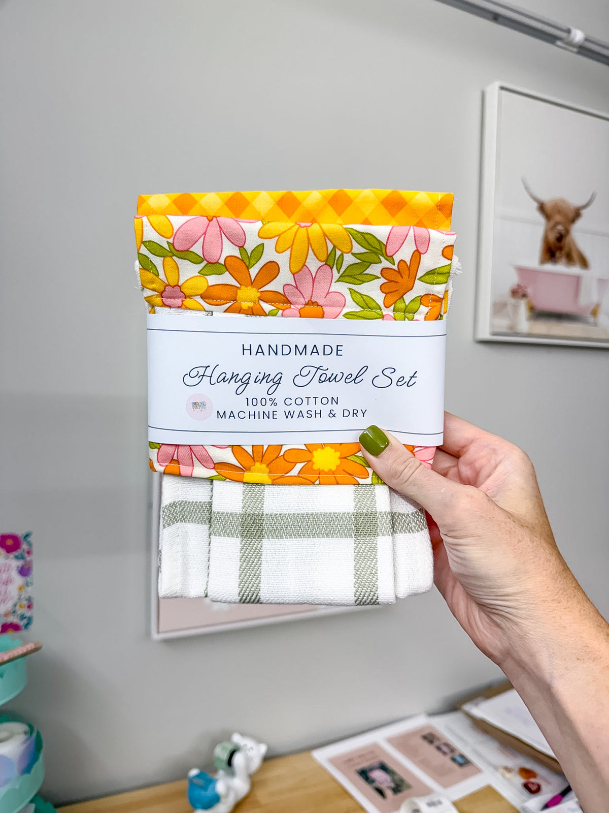 A hand holds a packaged hanging towel set against a gray wall. The set includes a folded floral towel with pink, orange, and yellow flowers, a folded orange-and-white floral towel, and a folded white towel with a sage green plaid pattern.