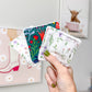 A hand with olive green nail polish holds a fan of four square fabric coasters with fringed edges. The coasters feature different floral and botanical patterns in green, red, purple, and white.