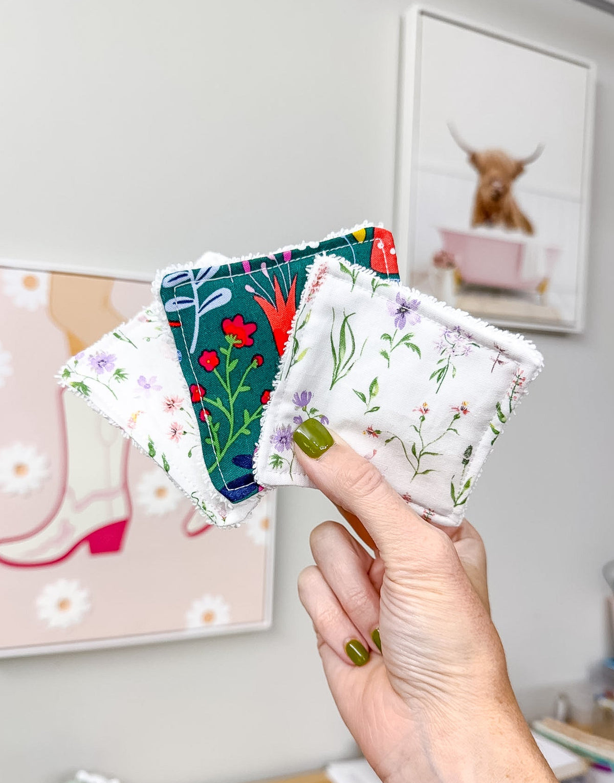 A hand with olive green nail polish holds a fan of four square fabric coasters with fringed edges. The coasters feature different floral and botanical patterns in green, red, purple, and white.