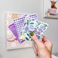 A hand holds three small square fabric coasters with different patterns: a purple gingham, a white floral print, and a purple floral print, against a light gray wall with framed art in the background.