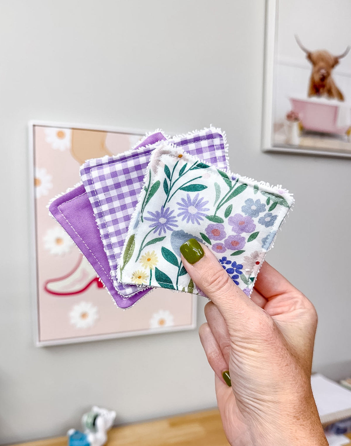 A hand holds three small square fabric coasters with different patterns: a purple gingham, a white floral print, and a purple floral print, against a light gray wall with framed art in the background.