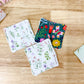 Three square reusable cloth napkins with stitched edges are arranged on a light wood tabletop. Two napkins have a white background with delicate green and purple floral prints, and the third napkin has a dark teal background with a colorful floral and bird pattern.