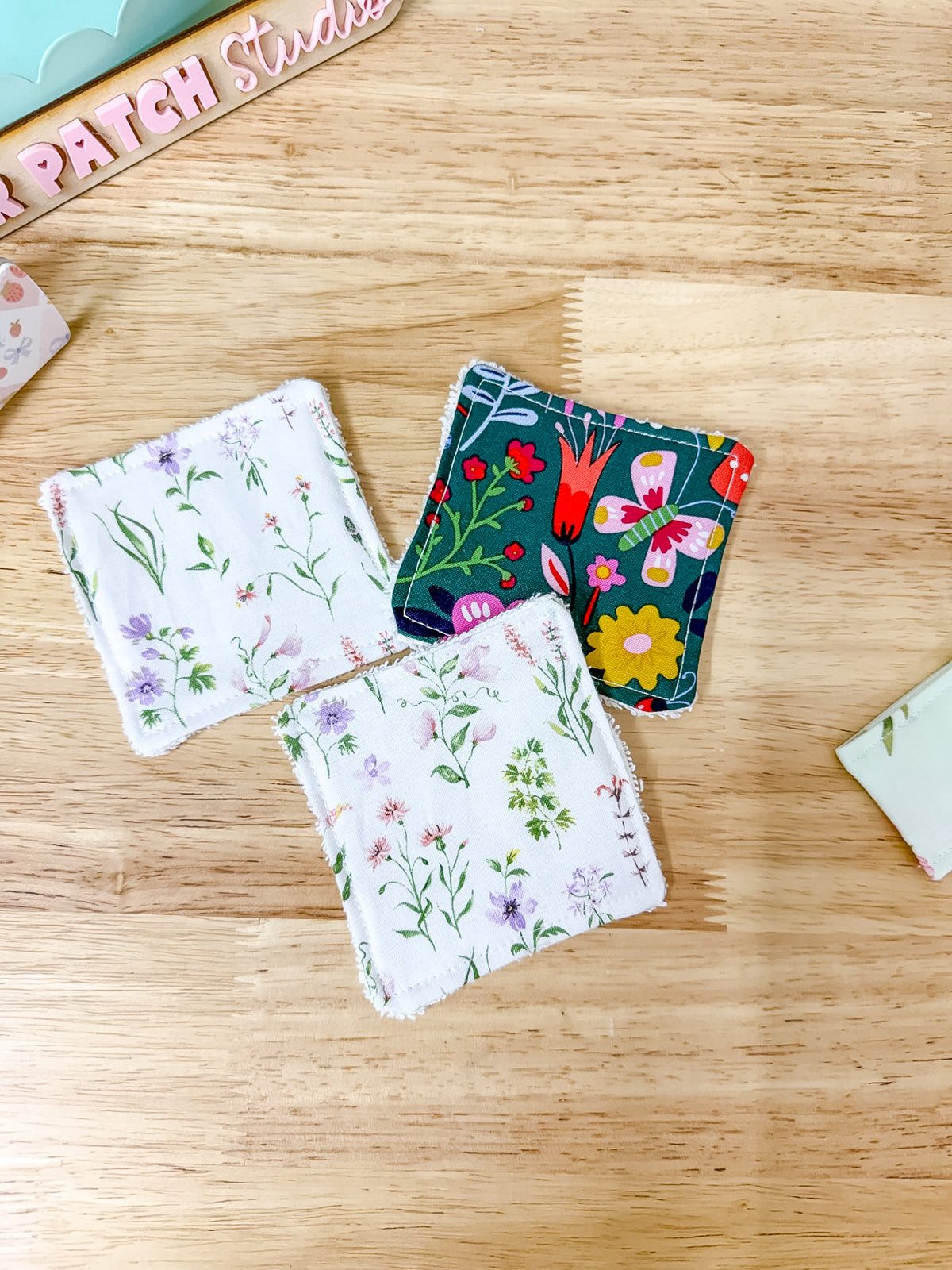 Three square reusable cloth napkins with stitched edges are arranged on a light wood tabletop. Two napkins have a white background with delicate green and purple floral prints, and the third napkin has a dark teal background with a colorful floral and bird pattern.