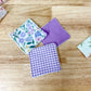 Three small fabric square patches with frayed edges are arranged on a light wood surface. One patch has a floral print in pastel colors, one is solid lavender purple, and one is a purple-and-white gingham pattern.
