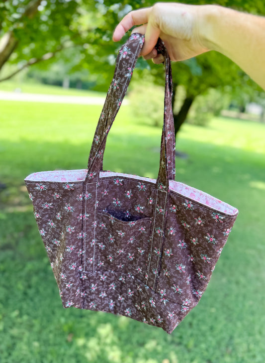 Person holding a floral-patterned tote bag outdoors with green grass and trees in the background