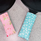 Two fabric pan handles covers resting on a gray textured surface, one pink with a red and green floral print and one light blue with white daisy patterns, next to black cast iron skillets.