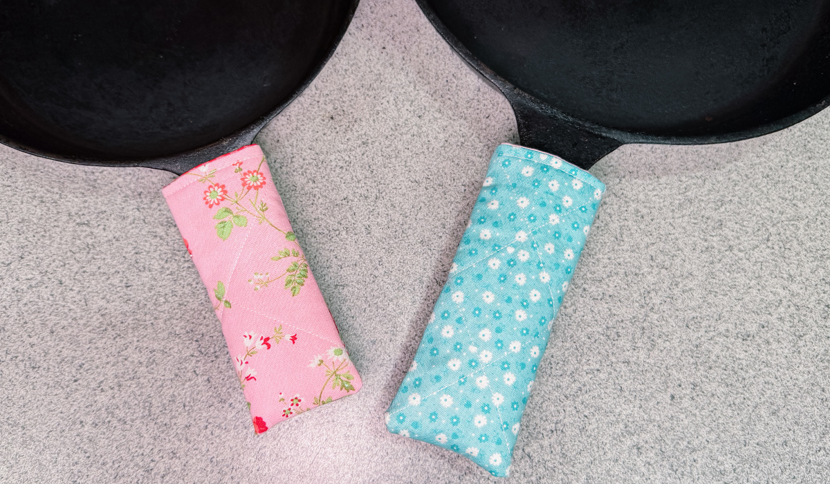 Two fabric pan handles covers resting on a gray textured surface, one pink with a red and green floral print and one light blue with white daisy patterns, next to black cast iron skillets.